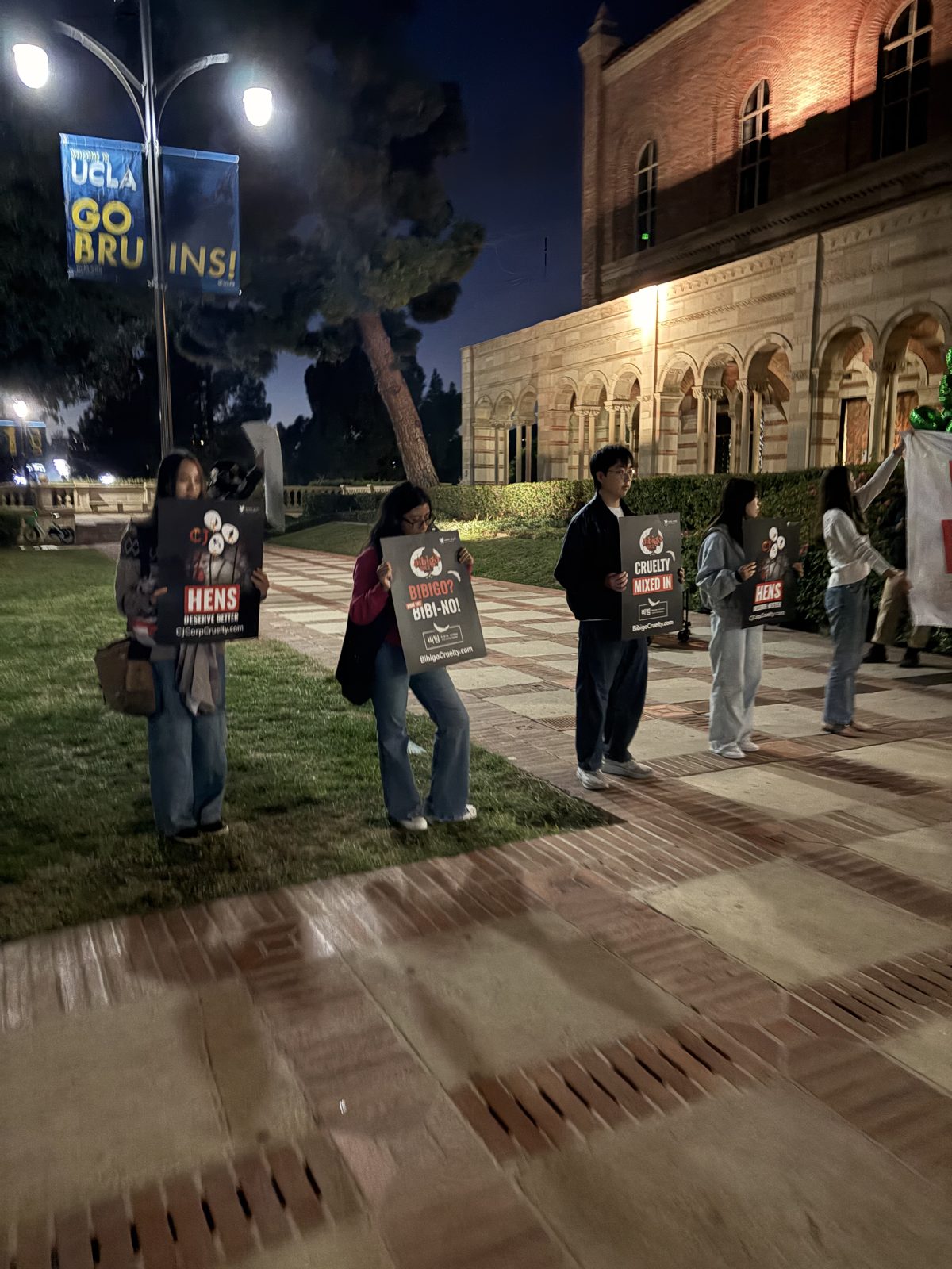 Students holding Bibigo cruelty protest signs outside Royce Hall at night